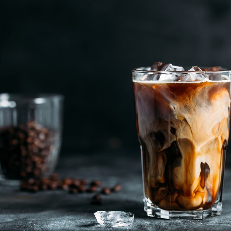 Ice coffee in a tall glass with cream poured over and coffee beans on dark gray background