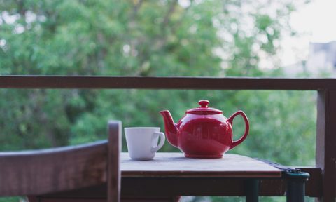 red teapot and white cup on wooden table outside with green trees in background