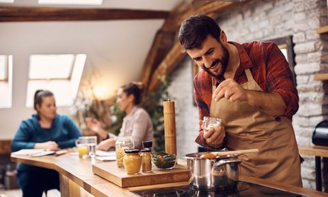 Happy man adding salt while cooking for his friends in the kitchen.
