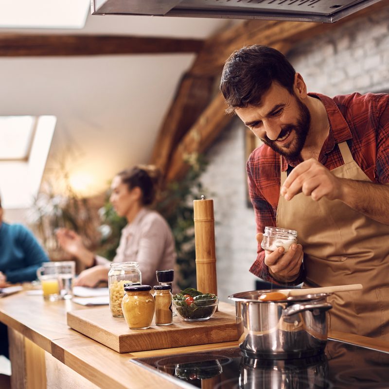 Happy man adding salt while cooking for his friends in the kitchen.