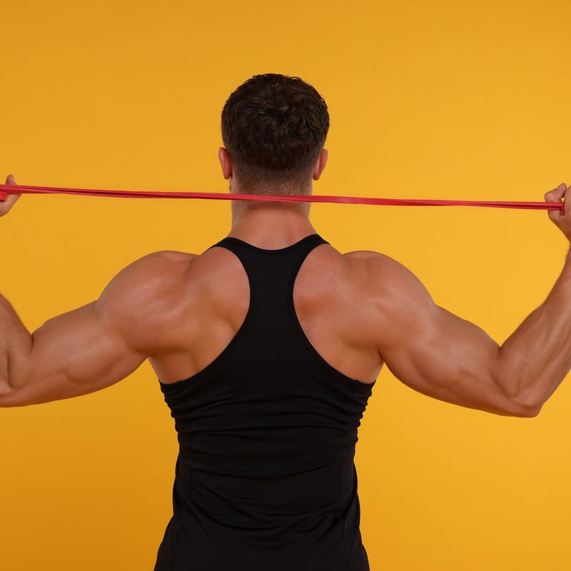 Young man exercising with elastic resistance band on orange background, back view