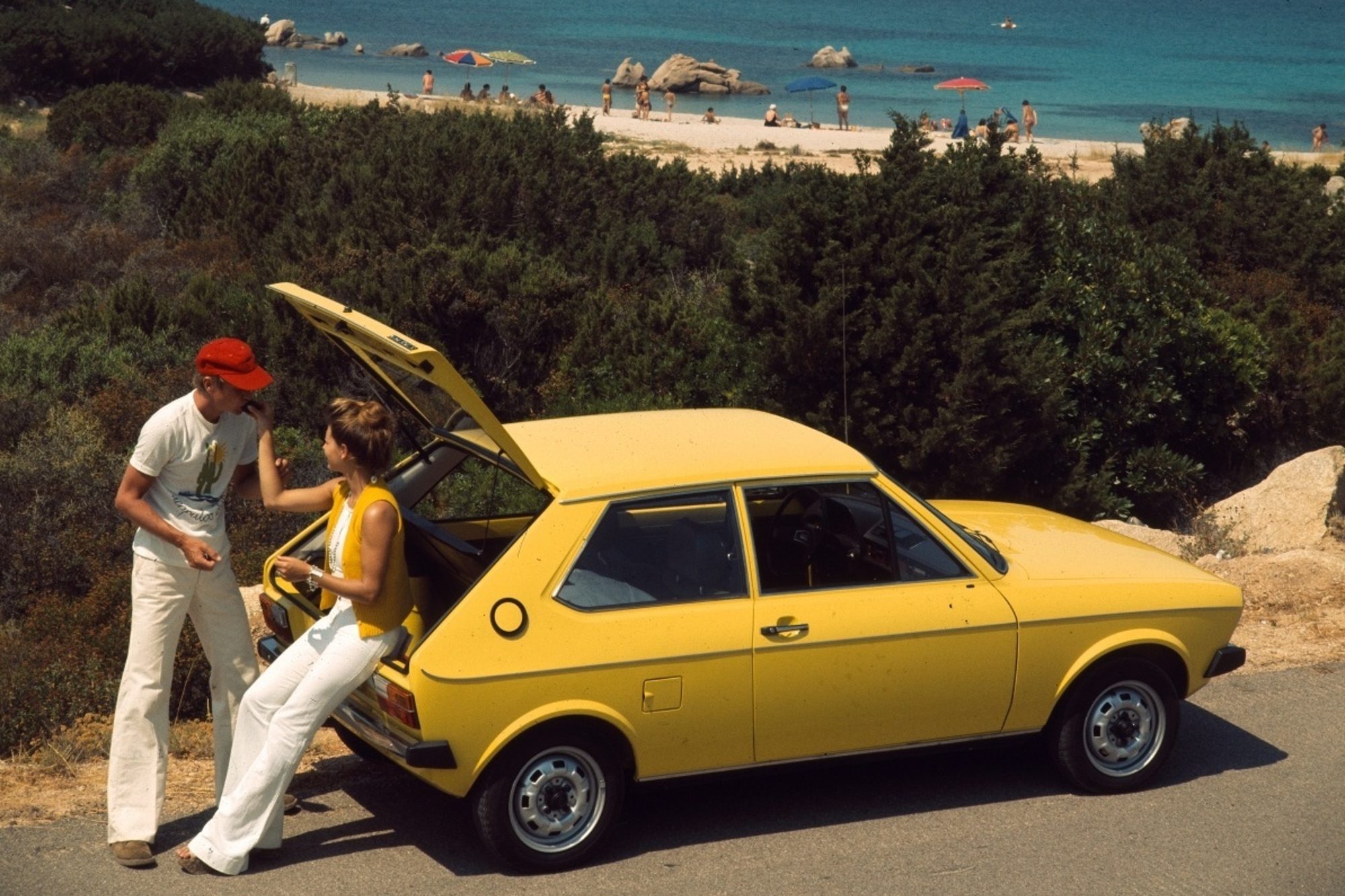 A couple of people feeding each other grapes from the back of an Audi 50 parked by the side of a road overlooking a beach.