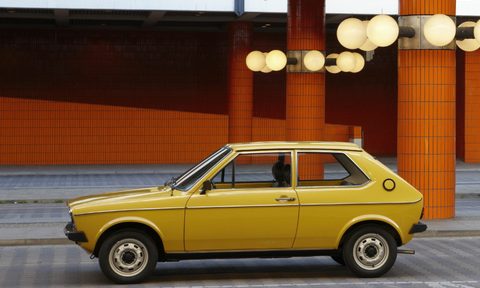 A yellow Audi 50 pared on the side of a roadway with a large red concrete building in the background.