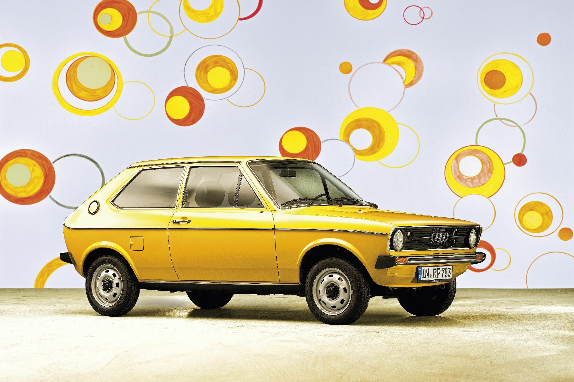A yellow Audi 50 parked in front of a backdrop wall with yellow and orange circles.
