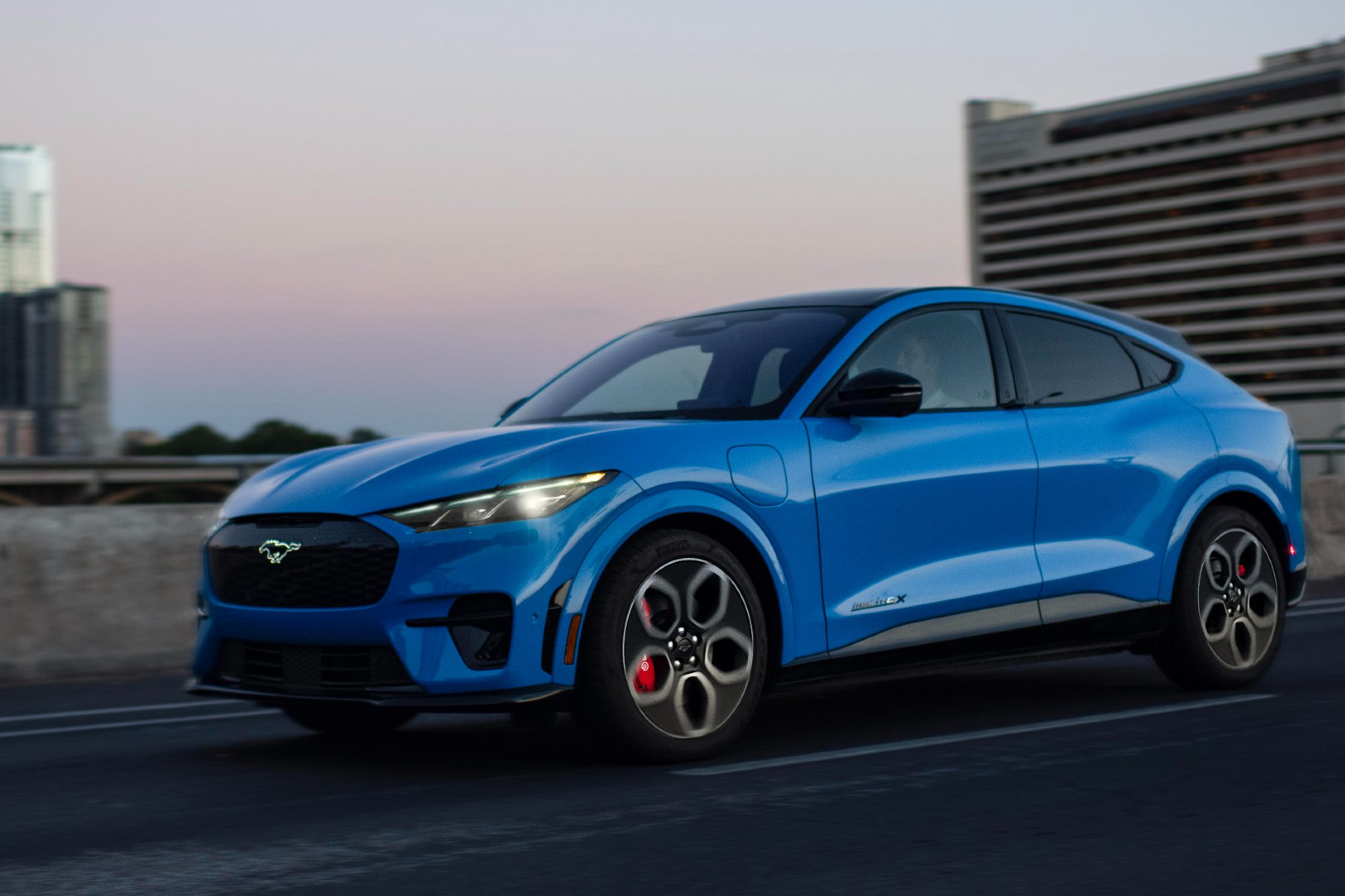 Blue 2024 Mustang Mach-E left front three-quarter shot traveling on a highway with large city commercial buildings in the background,