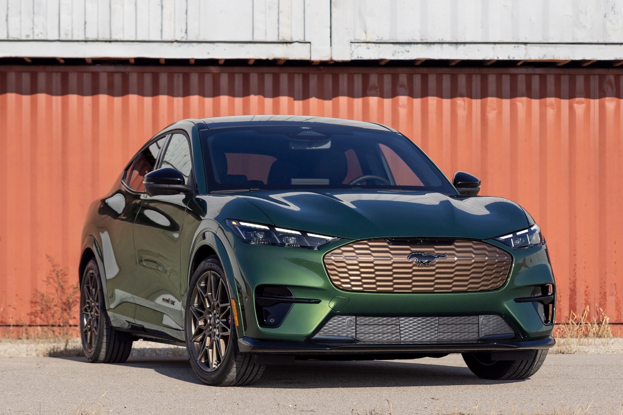 Green and bronze 2024 Ford Mustang Mach-E Bronze right front three-quarter view parked on gray sand in front of a dull red-ish color building.