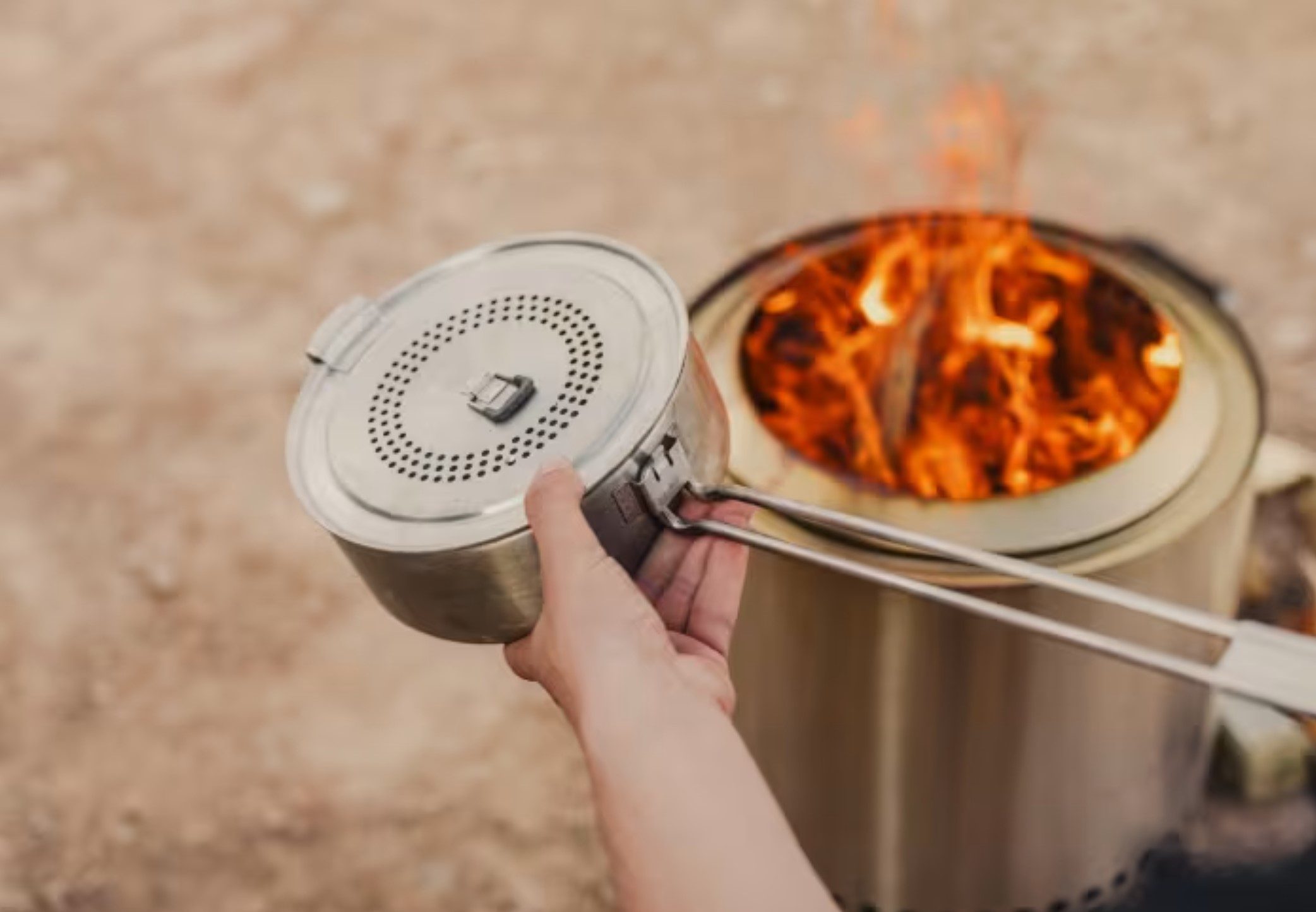 A person holding a popcorn maker over a fire pit