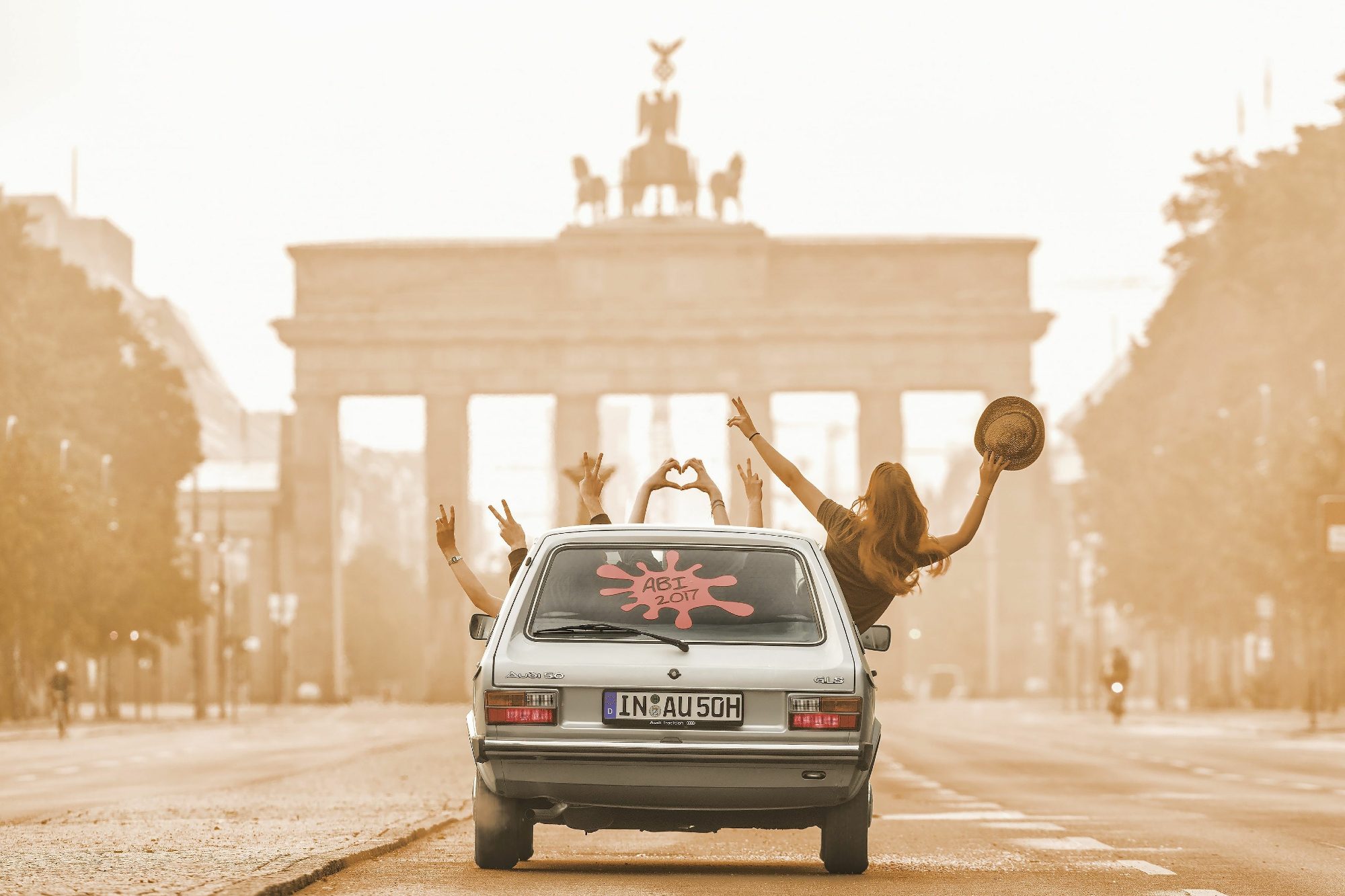 Light colored Audi 50 driving toward the Brandenburg Gate monument in Germany with passengers waving and one passenger leaning out of the window holding a hat.