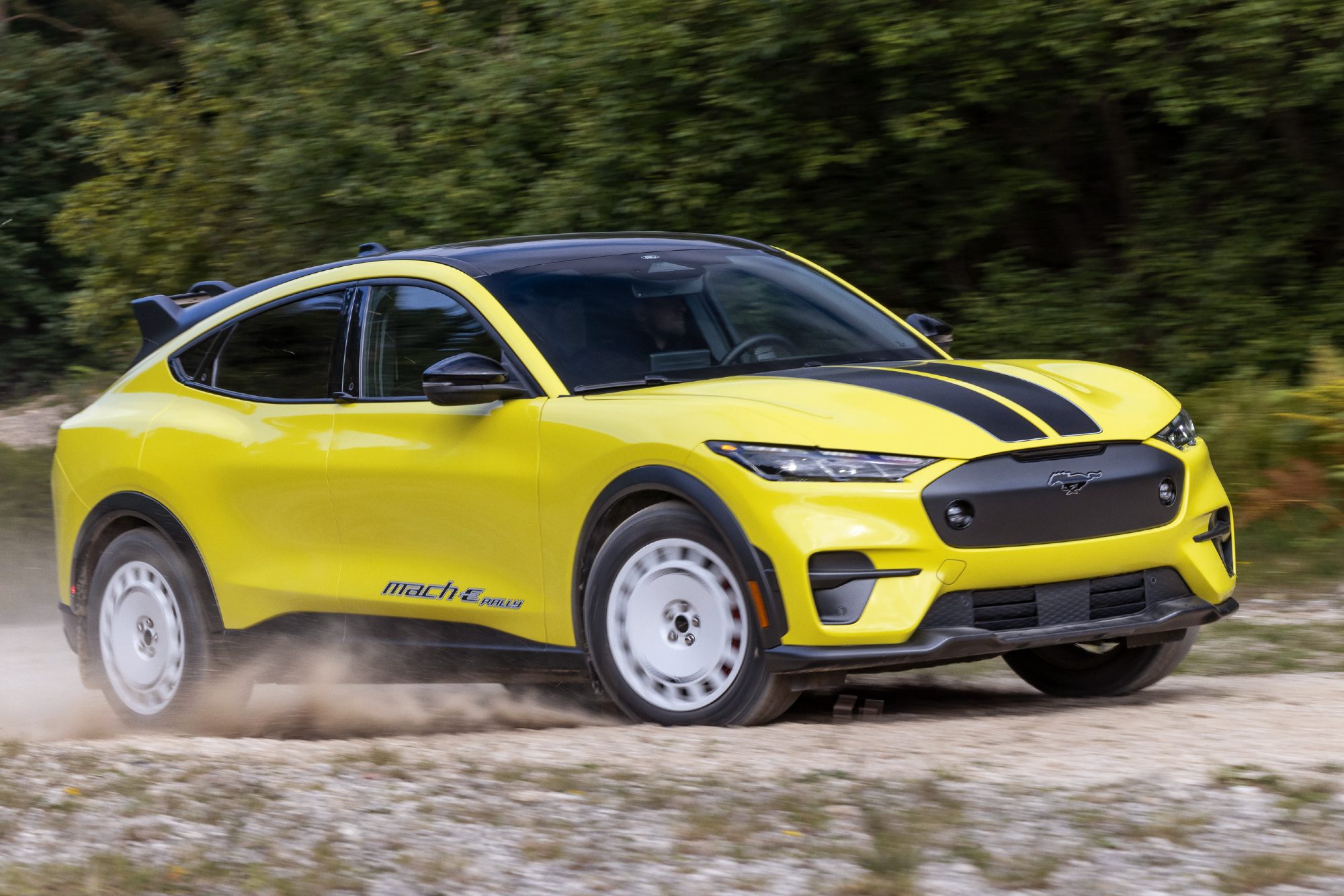 Yellow 2024 Mustang Mach-E Rally with black hood strips right front three-quarter view driving on dirt road with trees in the background.
