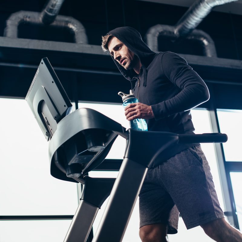 low angle view of handsome sportsman in hoodie exercising on treadmill and holding sport bottle with water in gym