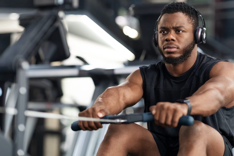 Closeup of handsome african american sportsman training body on modern rowing exerciser in gym, copy space. Motivated young black sportsman with wireless headset having workout on rowing machine