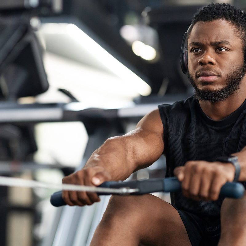 Closeup of handsome african american sportsman training body on modern rowing exerciser in gym, copy space. Motivated young black sportsman with wireless headset having workout on rowing machine