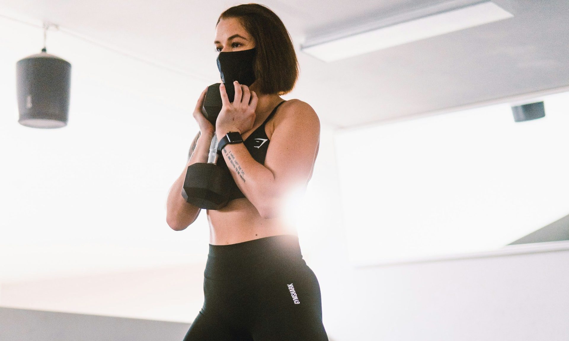 woman wearing mask ready to goblet squat holding weight dumbbell wearing leggings in gym