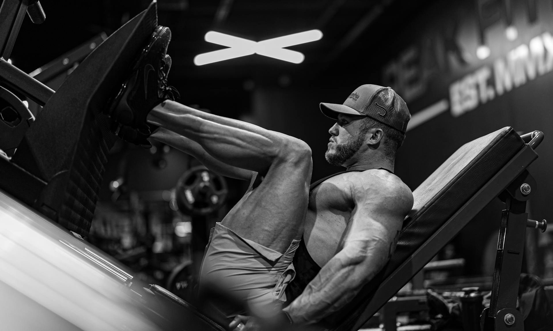 black and white image of man in gym wearing hat and shorts performing leg press exericse on machine