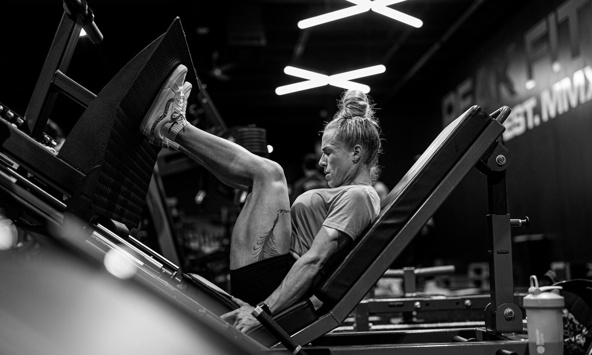 black and white image of woman wearing t shirt and shorts in gym performing leg press on machine