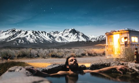 A man soaking in a natural tub outside of an RV during dusk with a mountain range behind him.