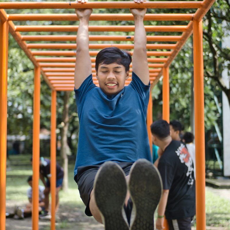 Man wearing blue shirt outside hanging on monkey bars smiling doing hanging hold exercise