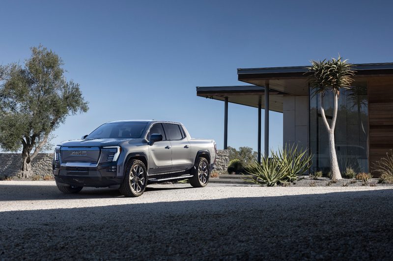 2024 GMC Sierra EV Denali Edition 1 parked on a crushed rock driveway in front of a single story, mountain home.