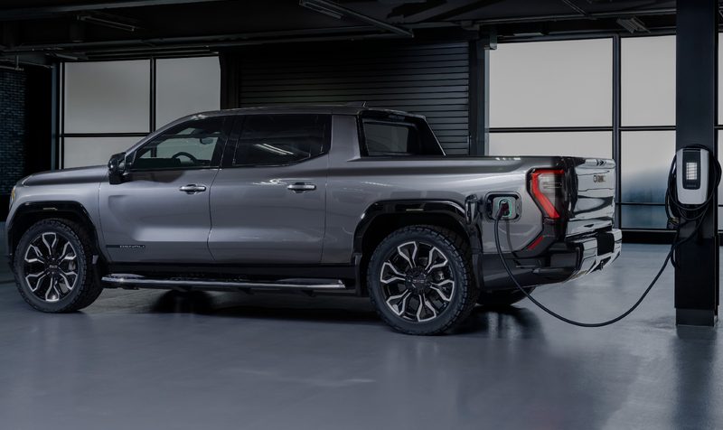 2024 GMC Sierra EV Denali Edition 1 plugged into a Level 2 charger inside an otherwise vacant area with concrete floor and concrete and glass wall.