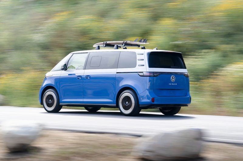 2025 VW ID. Buzz with a surfboard on a roof rack traveling on a road near a beach.