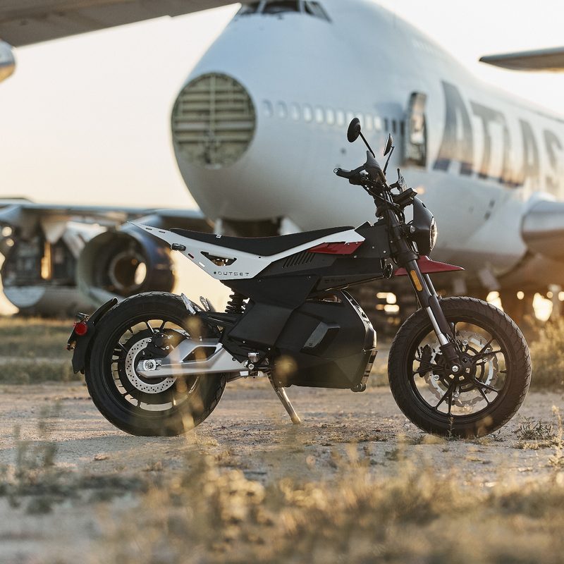 A Sector Red Ryvid Outset electric motorcycle right profile shot parked in desert among huge parked and out-of-service airplanes.