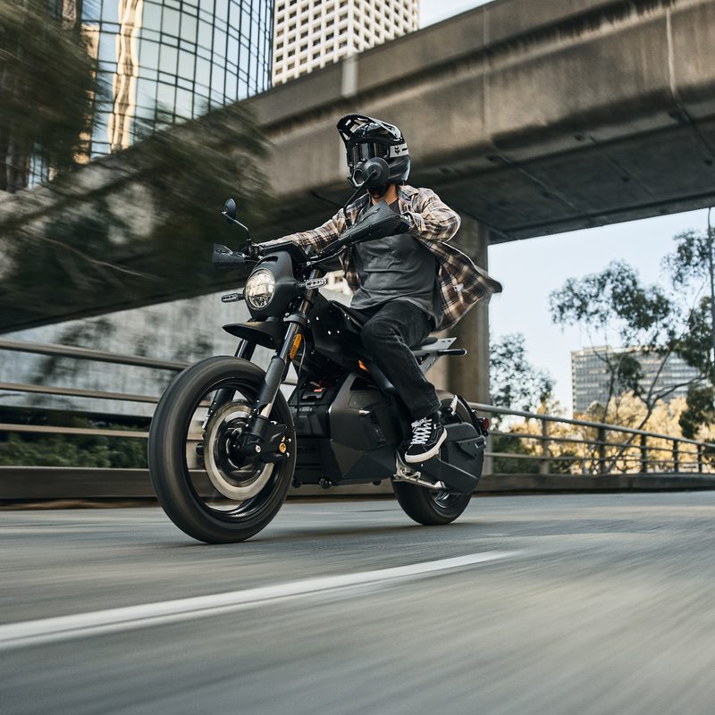 A rider on a Ryvid Outset electric motorcycle in Vapor Grey on a city street under an overpass.