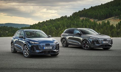Audi Q6 e-tron quattro and Aui Q6 e-tron parked on a gray sand surface with forested hills and mountains in the background.