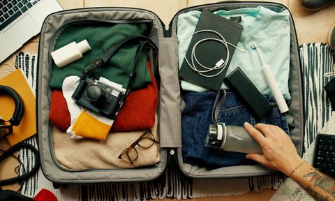 Man with tatted arm packs suitcase for adventure travel trip. Packing bag with camera, laptop, portable battery charger and electric toothbrush.