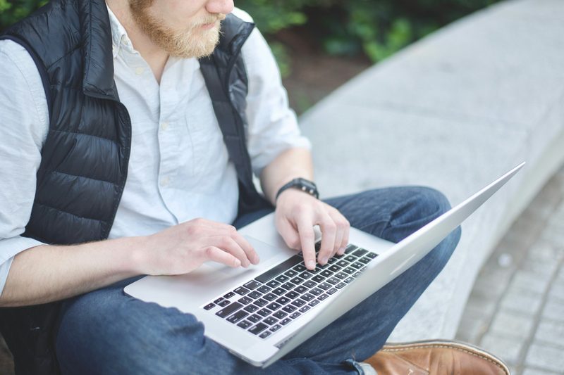 A man sitting on a bench outside while typing on a laptop