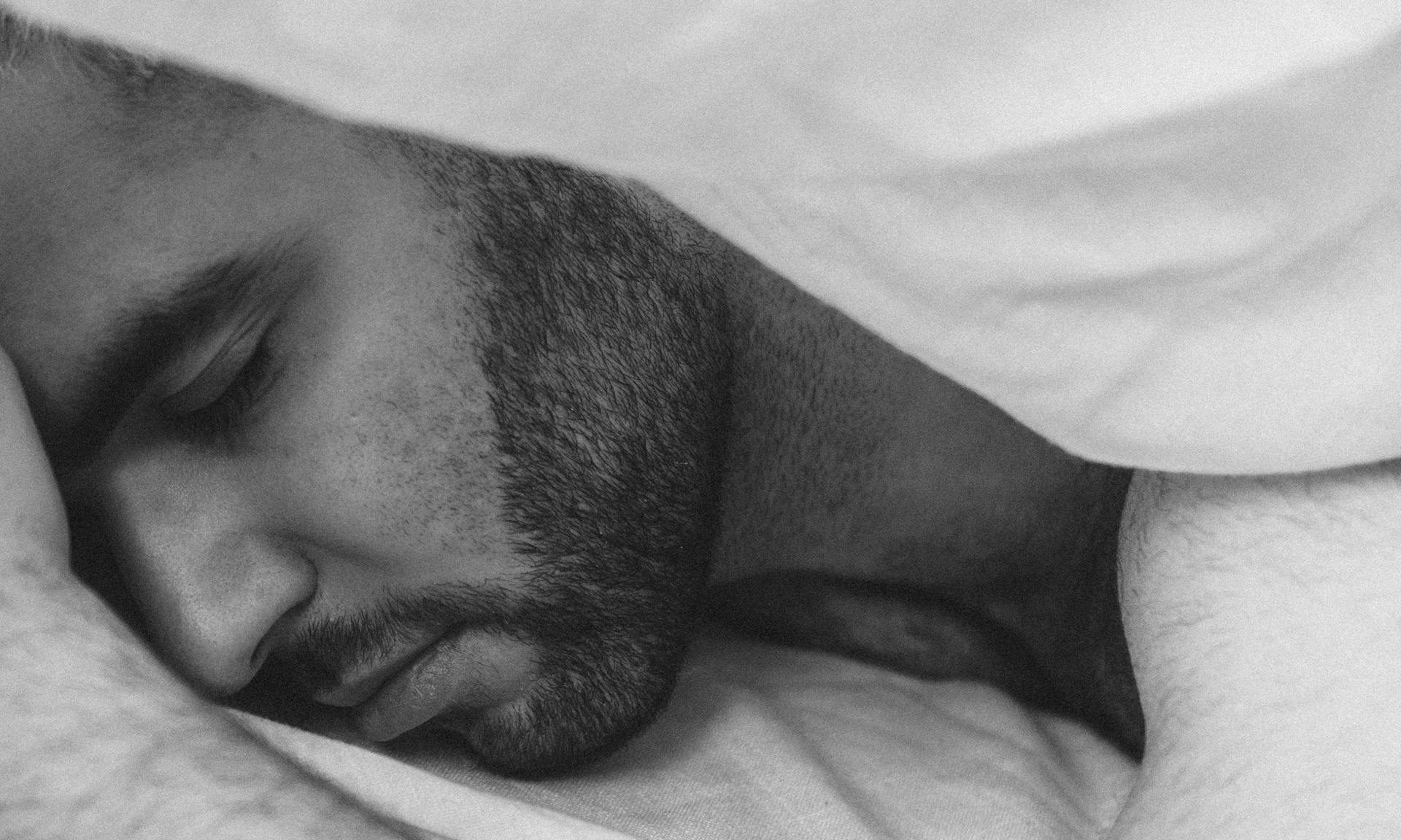 man sleeping in bed under duvet with beard black and white image