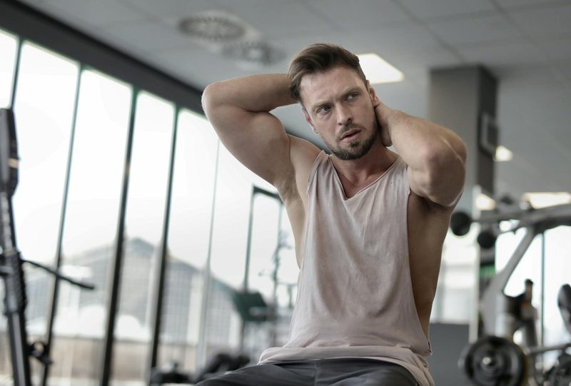 Man sitting in gym wearing vest or tank top flexing biceps bench tank