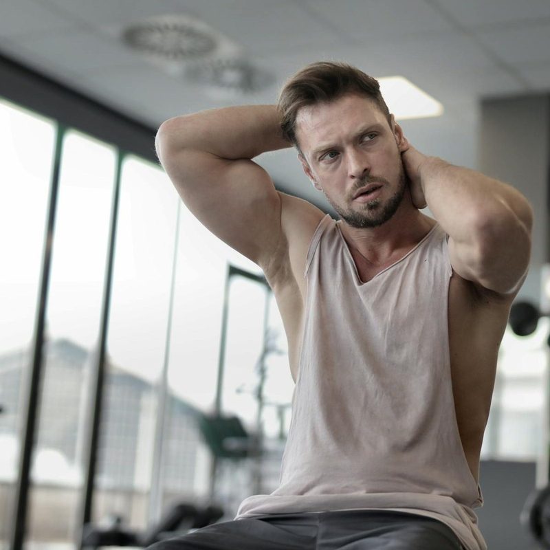 Man sitting in gym wearing vest or tank top flexing biceps bench tank