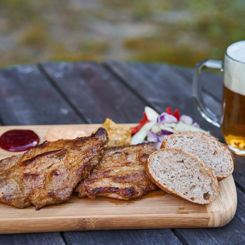 Pork collar steaks served with slices of whole grain bread, choped onions, tomatoes, mustard and barbeque sauces on the wooden plate outside with mug of pilsner czech beer.
