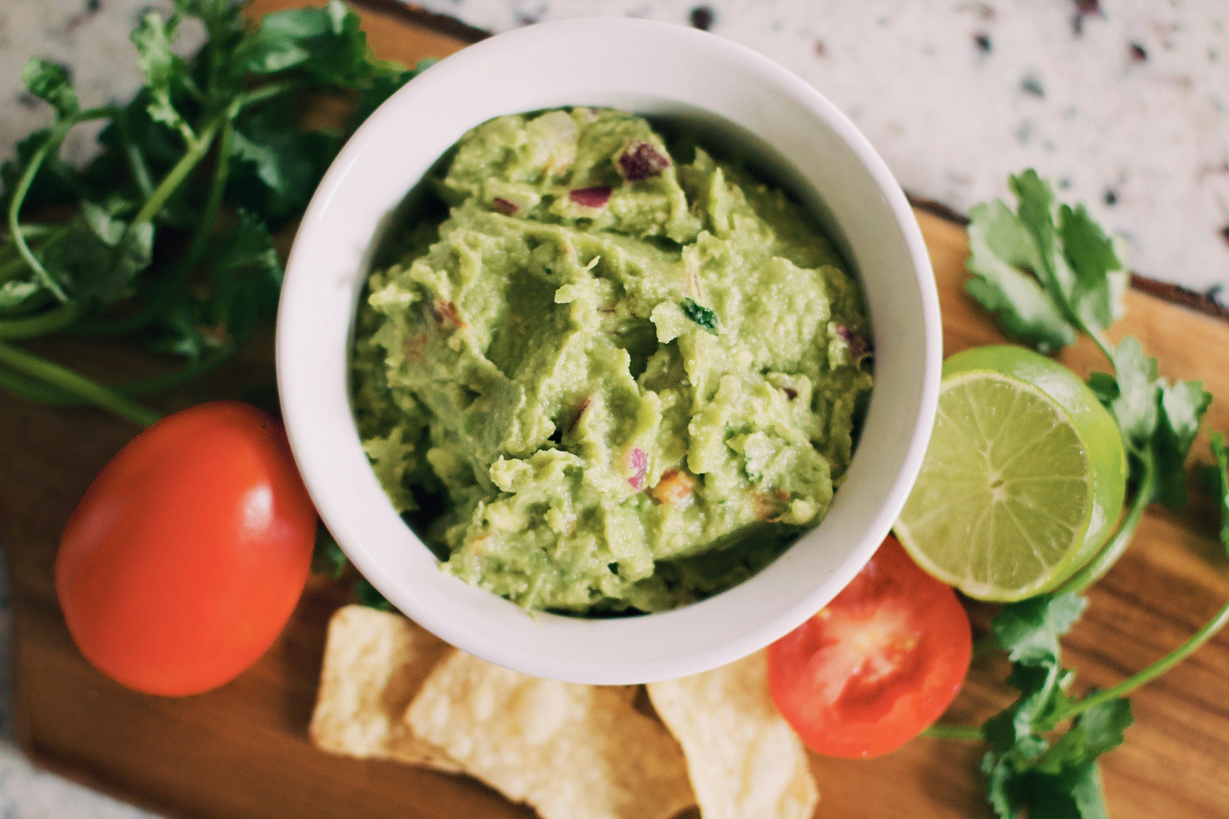 guacamole in white bowl with herbs and tomatoes on wooden counter
