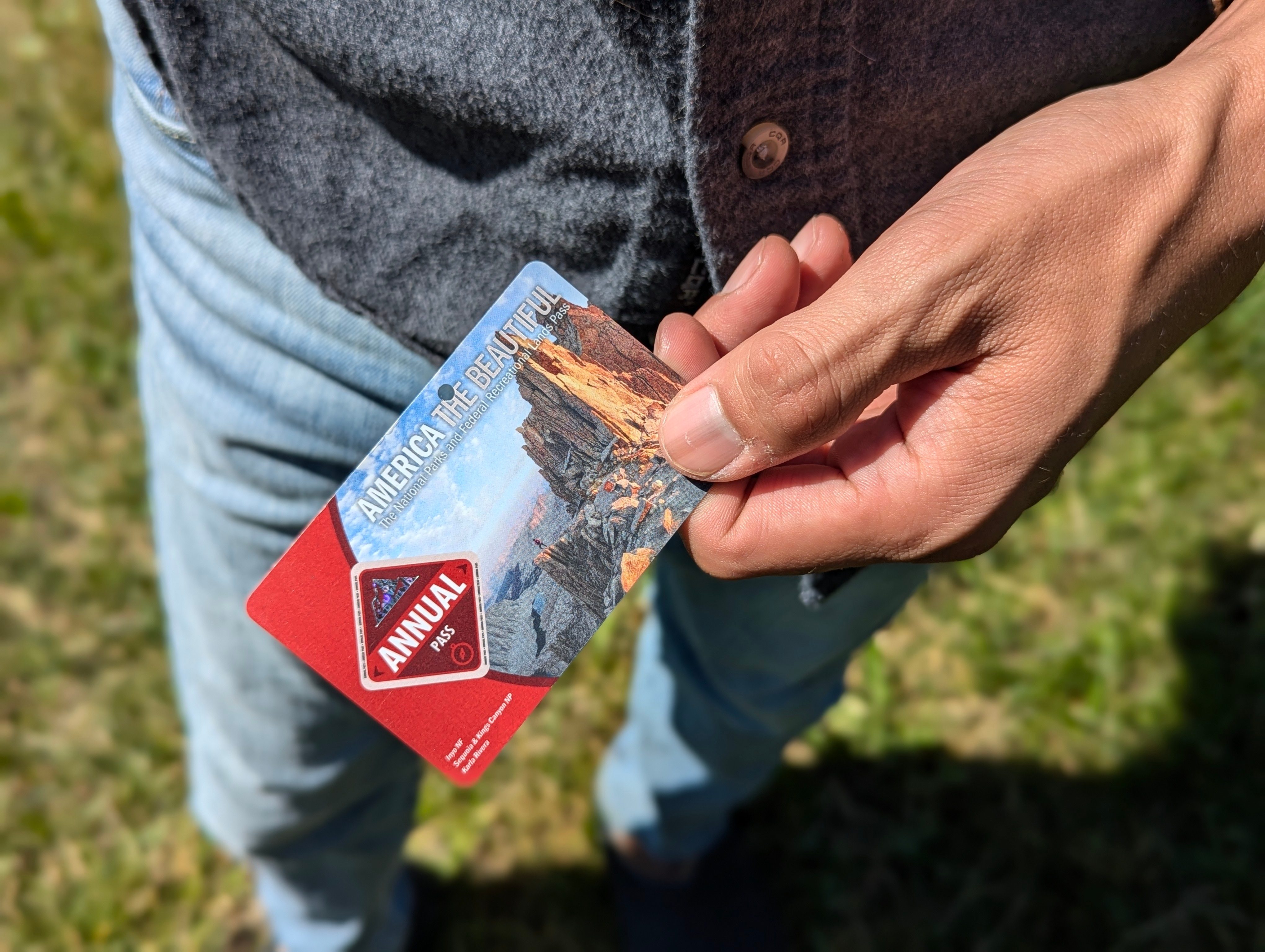 A man holding an America the beautiful parks pass wearing a button down top and blue jeans standing outdoors