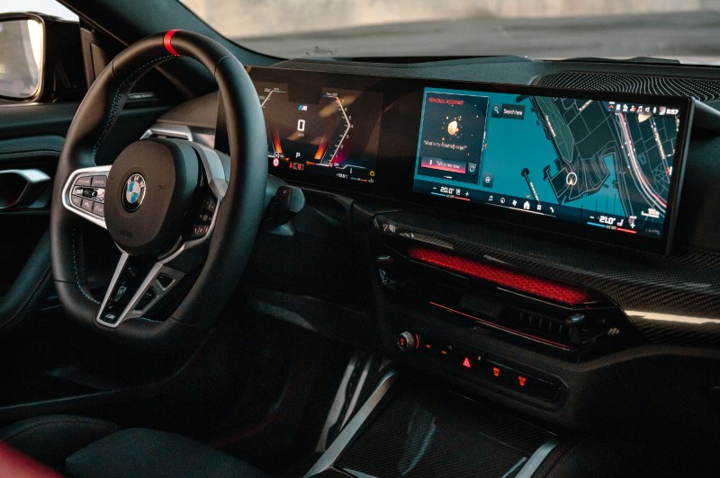 2025 BMW 2 Series Coupe interior view from posiiton of front passenger headrest looking toward the driver cockpit showing driver information and infotainment displays.