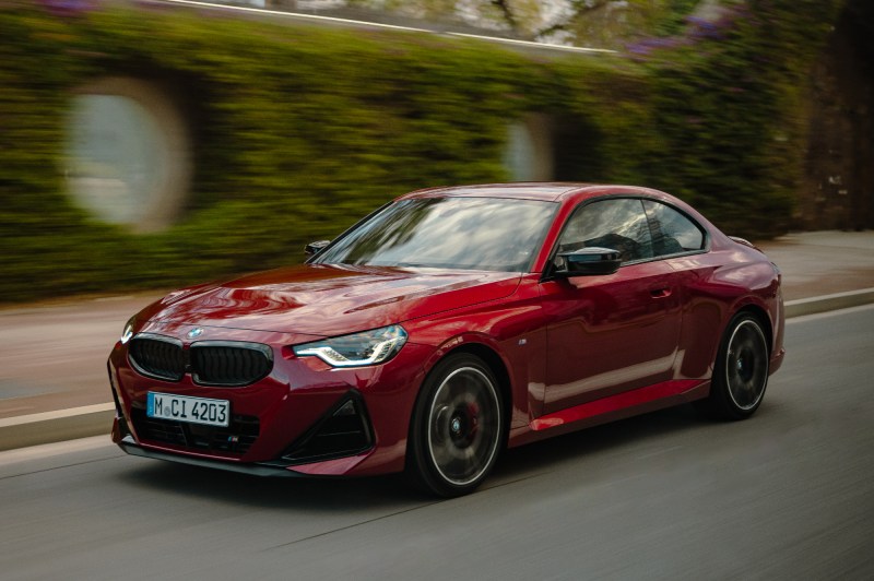 2025 BMW 2 Series Coupe left front three-quarter view driving on a street with a sidewalk and ivy-covered brick walls in the background.