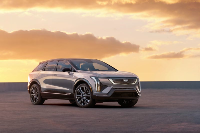2025 Cadillac Optiq right front three-quarter view parked on gray sand with a sunrise-filled sky in the background.