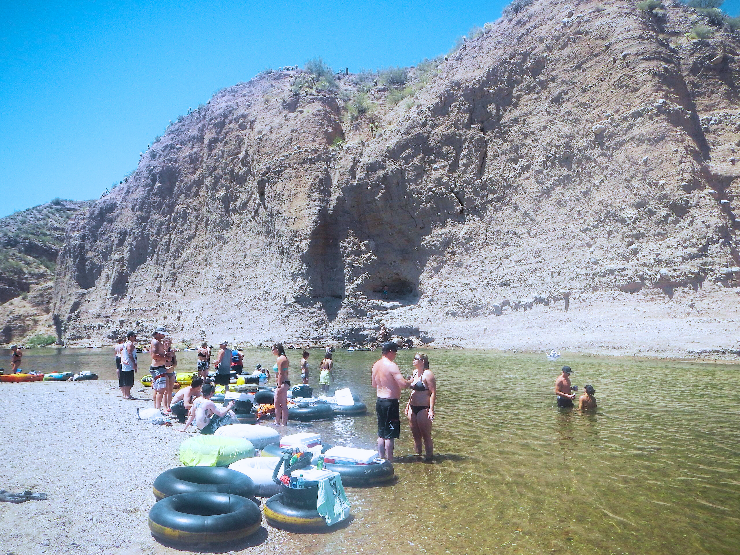 a group of outdoor enthusiasts floating down the salt river alongside a sandy bank on a sunny day