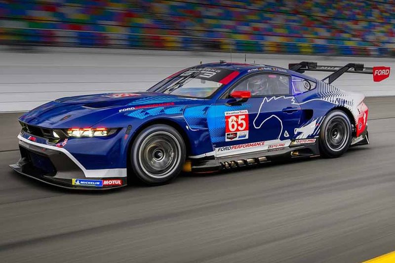 Ford Mustang GT3 race car ON race track rushing by a stadium full of fans.