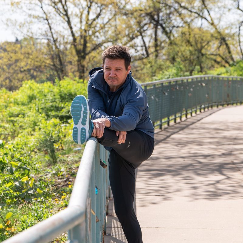 Jeremy Renner actor wearing brooks running shoes stretching outside recovery from injury