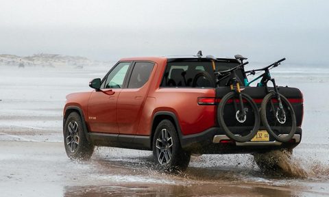 Red second-generation Rivian R1T right rear three-quarter view parked on beach edge facing water bike on back of the truck.