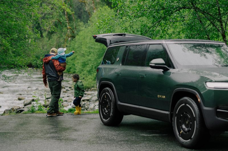 Second-generation Rivian R1S right front three-quarter view backed up to a stream with father holding one child with a second child standing