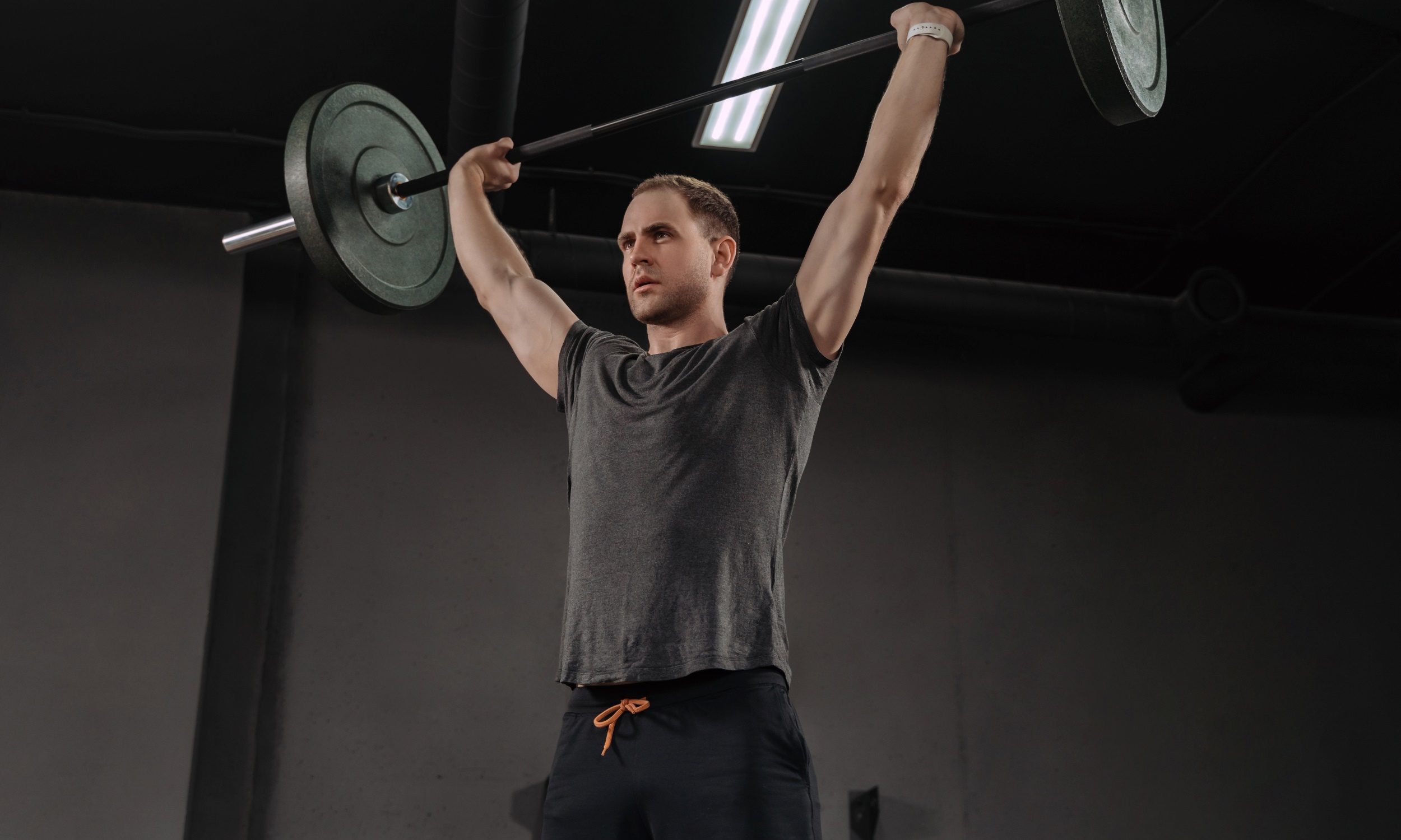 man wearing grey t shirt in gym doing overhead press or military press with barbell