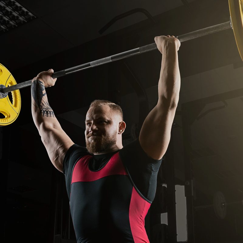 Man lifting overhead press military press exercise with barbell in gym wearing black and red shorts