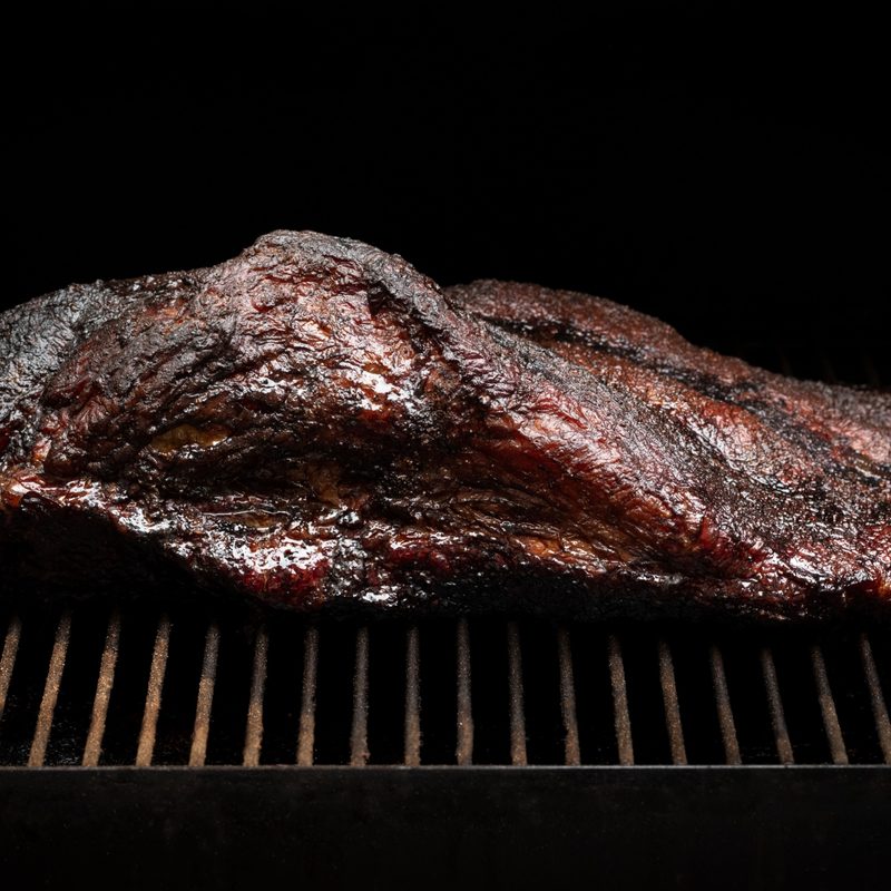 Slow cooked beef brisket on the grill grates of a smoker barbecue, in a cooking background