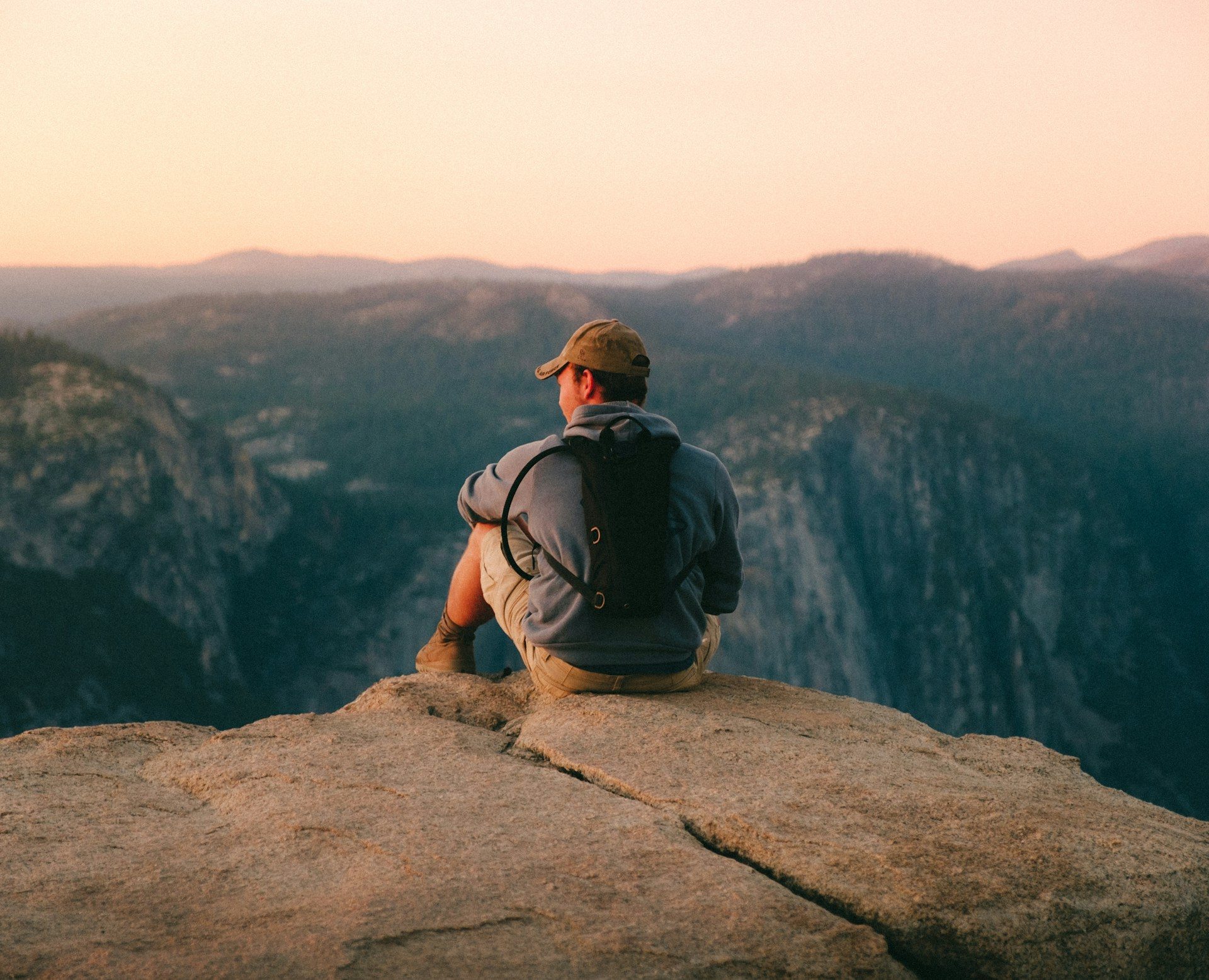 A young man sitting on a cliff in a natural setting during dusk or sunrise