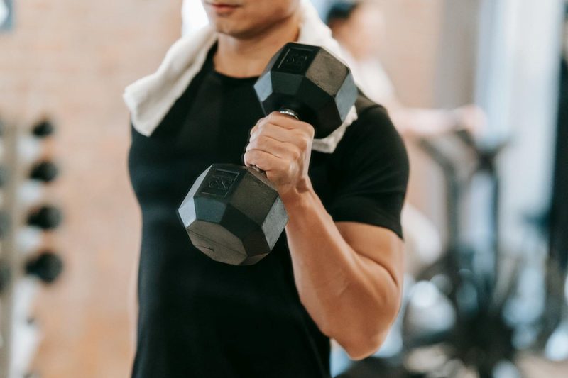 man wearing black T shirt at gym with towel around neck holding dumbell doing one arm exercise curl