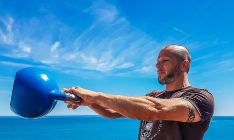 Man swinging blue kettlebell outside by blue ocean and blue sky