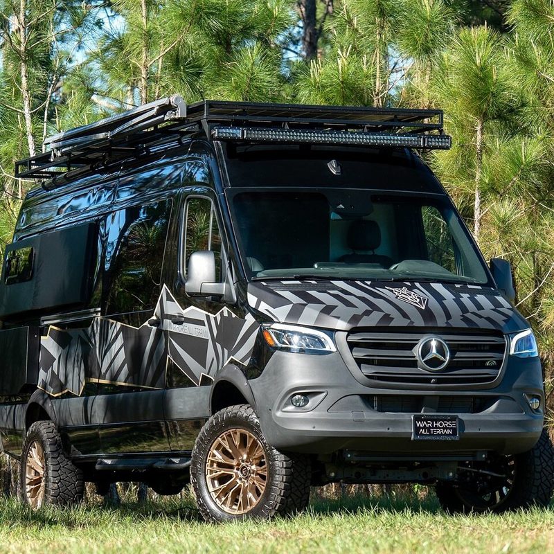 A black War Horse All-Terrain camper van parked among a stand of trees.